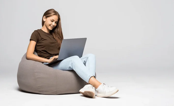 A teenage girl is sitting on a comforter, holding a laptop and wearing white sneakers.